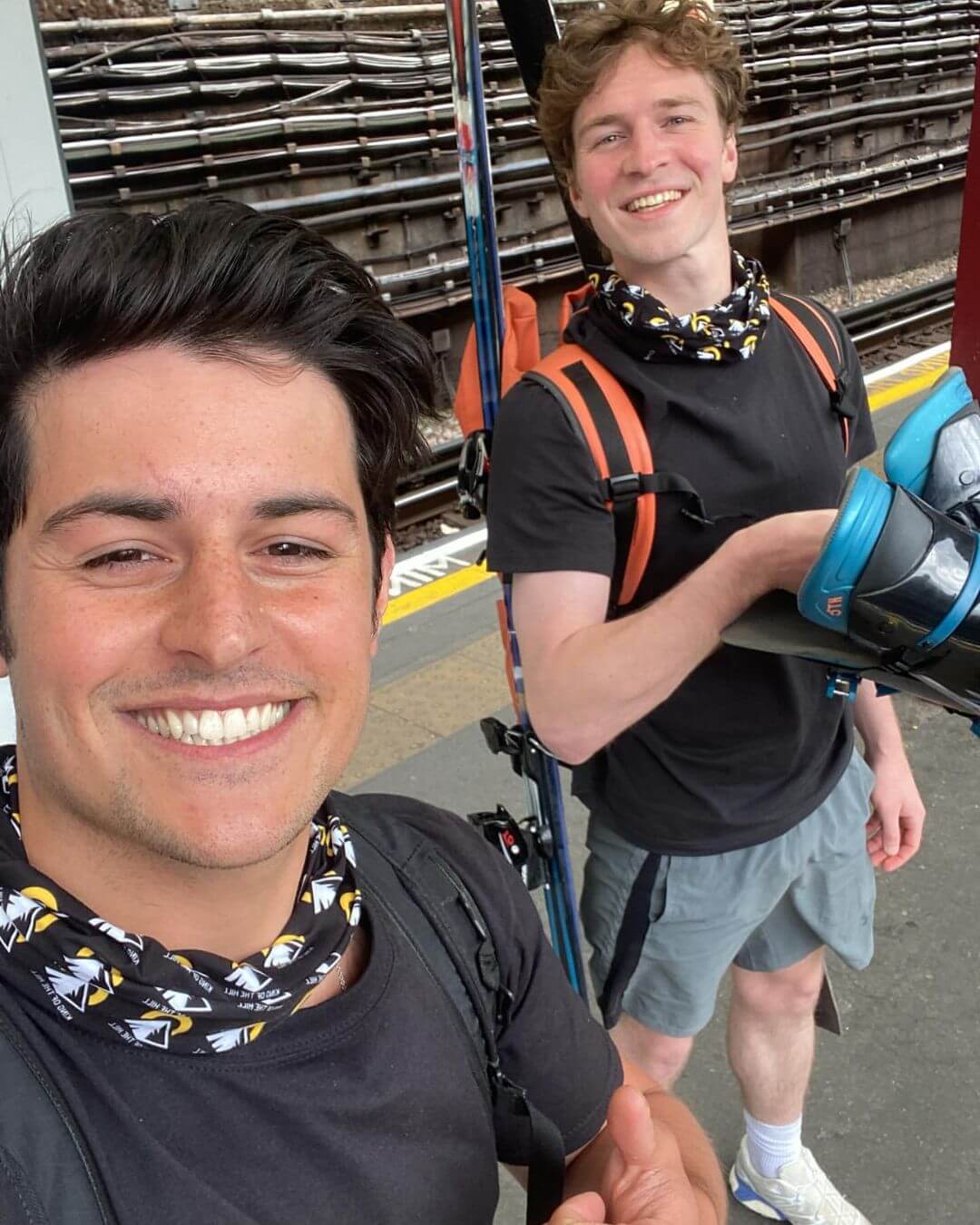 Matt and Reuben smile at a train station platform, wearing black King of the Hill neck gaiters and carrying skis as part of their snow camp fundraising journey from London to Val Thorens in ski boots.