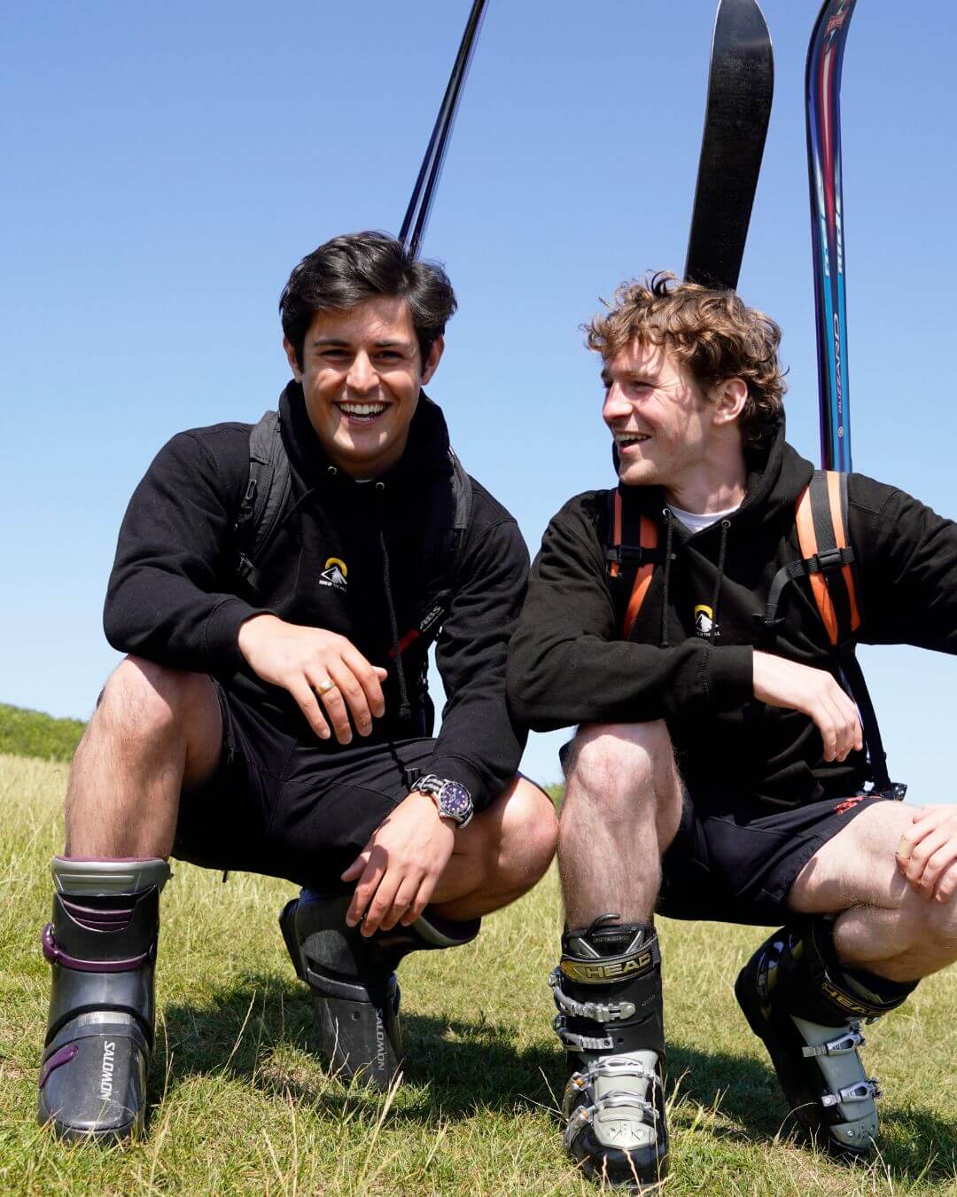 Two men, Matt and Reuben from King of the Hill, smile while kneeling on a grassy hilltop in full ski boots with skis strapped to their backpacks. They're mid-journey on a unique snow camp fundraising mission, walking from London to Val Thorens to support youth through Snow Camp UK.