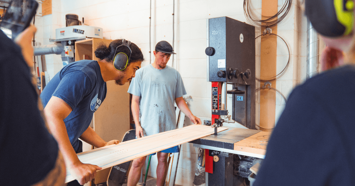 A young man wearing ear protection uses a bandsaw to cut a snowboard plank while another young person observes in a workshop, highlighting hands-on training in the Futures snow camp programme.