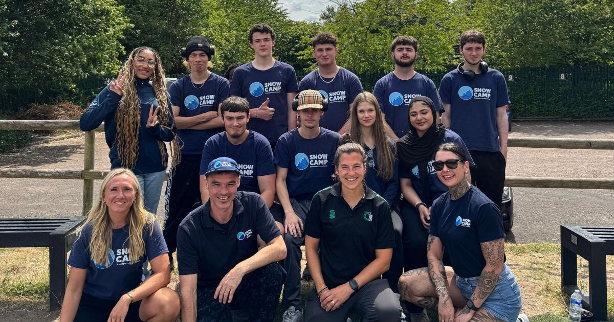 Group photo of a diverse group of young people and adults outdoors, many wearing navy blue "Snow Camp" T-shirts. They are smiling and posing together in front of a wooden fence and greenery, suggesting a team or program gathering in a casual, sunny setting.