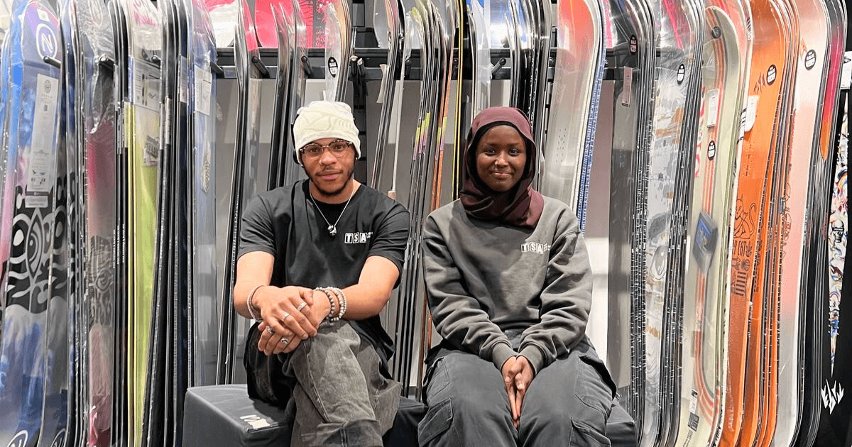 Two young adults sit between rows of snowboards in a shop, smiling and wearing matching branded shirts, showcasing retail and industry work experience opportunities through the Futures programme.