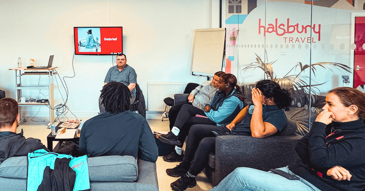 A group of young people sit in a casual meeting room listening to a man leading a debrief session with 'Halsbury Travel' branding in the background, representing team training and industry partnerships in the Futures snow camp.