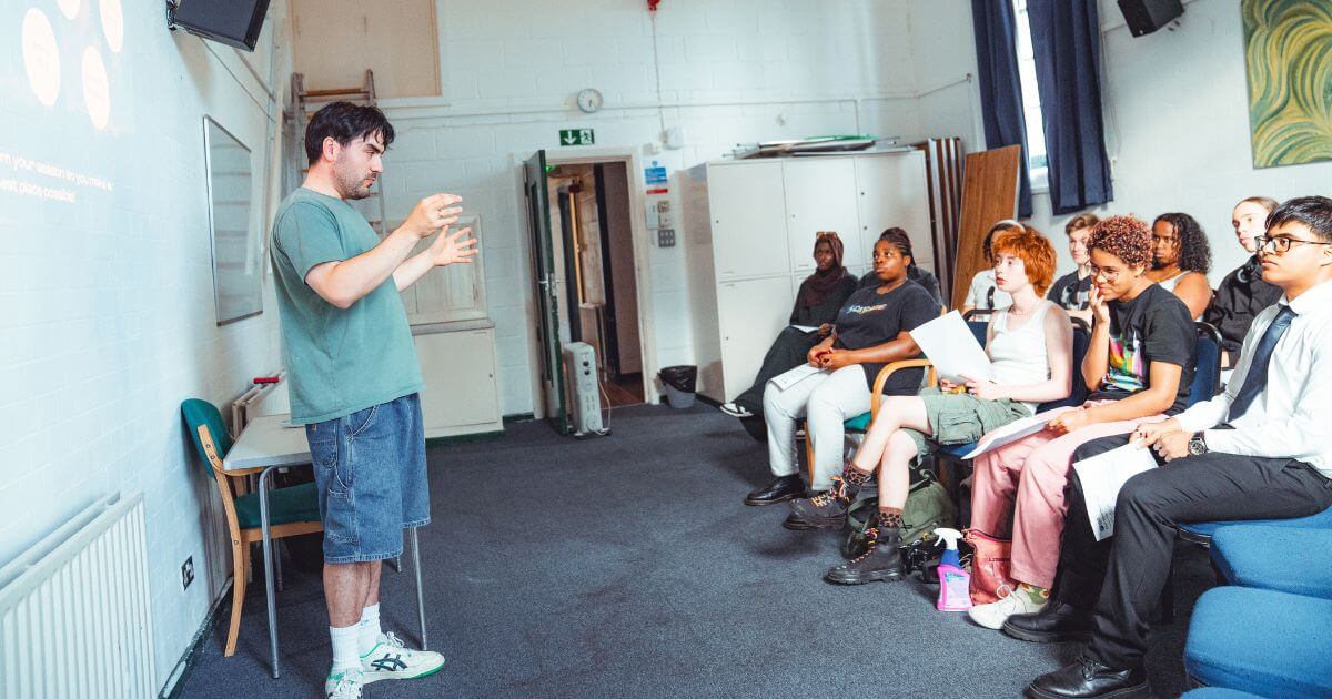 A young man gestures while speaking in front of a group of diverse young people seated in a classroom, holding papers and listening attentively as part of Futures programme workshops.