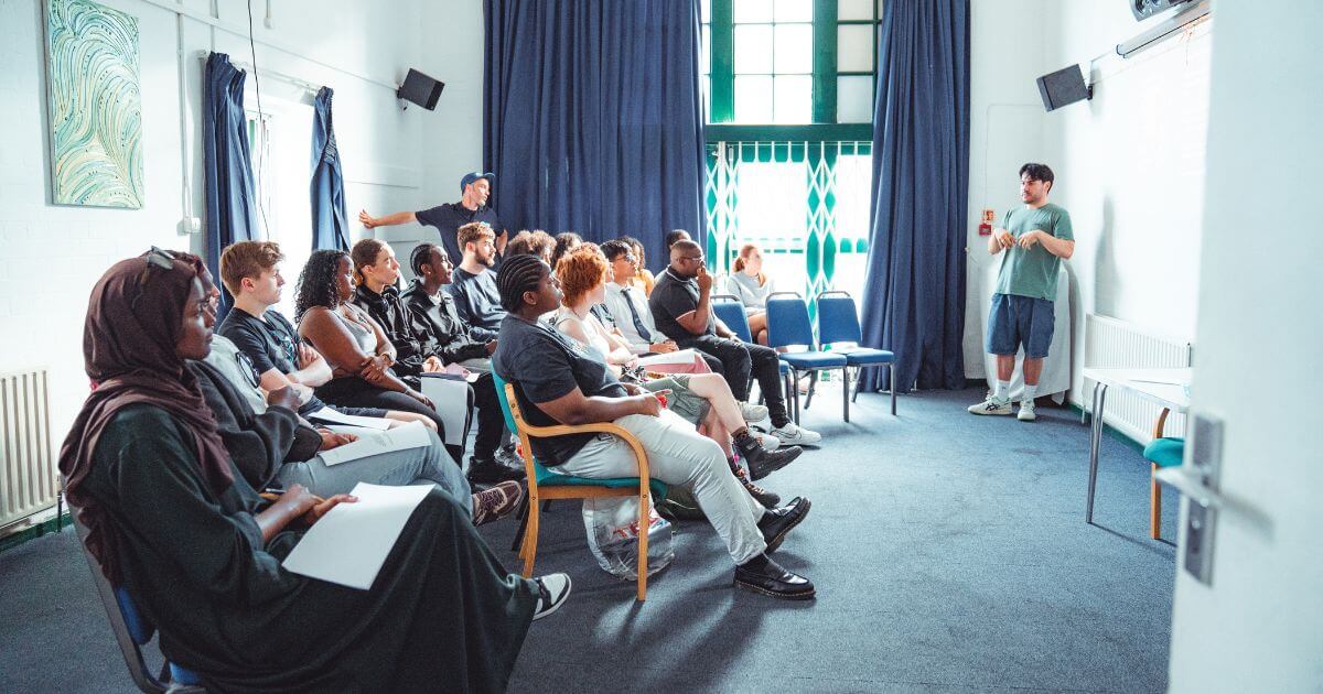 A diverse group of young adults listen attentively to a speaker at the front of a bright classroom with large windows and navy curtains. They appear engaged in a group training session.