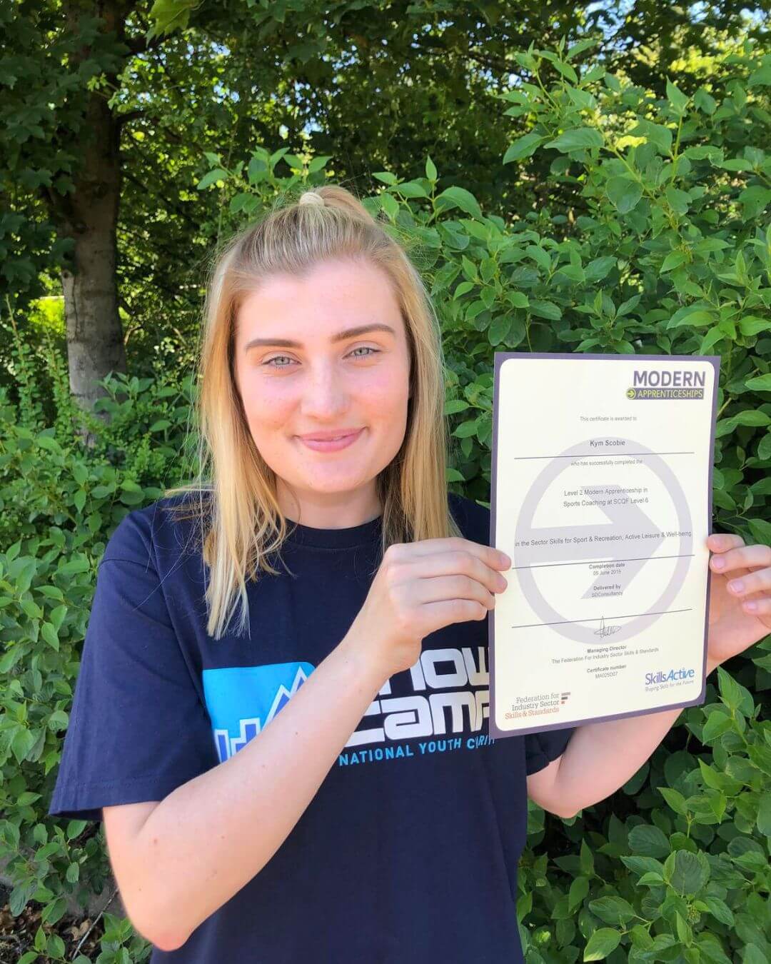 A young woman stands outdoors in front of leafy greenery, smiling while holding a certificate from the Modern Apprenticeships program in Sports Coaching. She wears a navy Snow Camp T-shirt.
