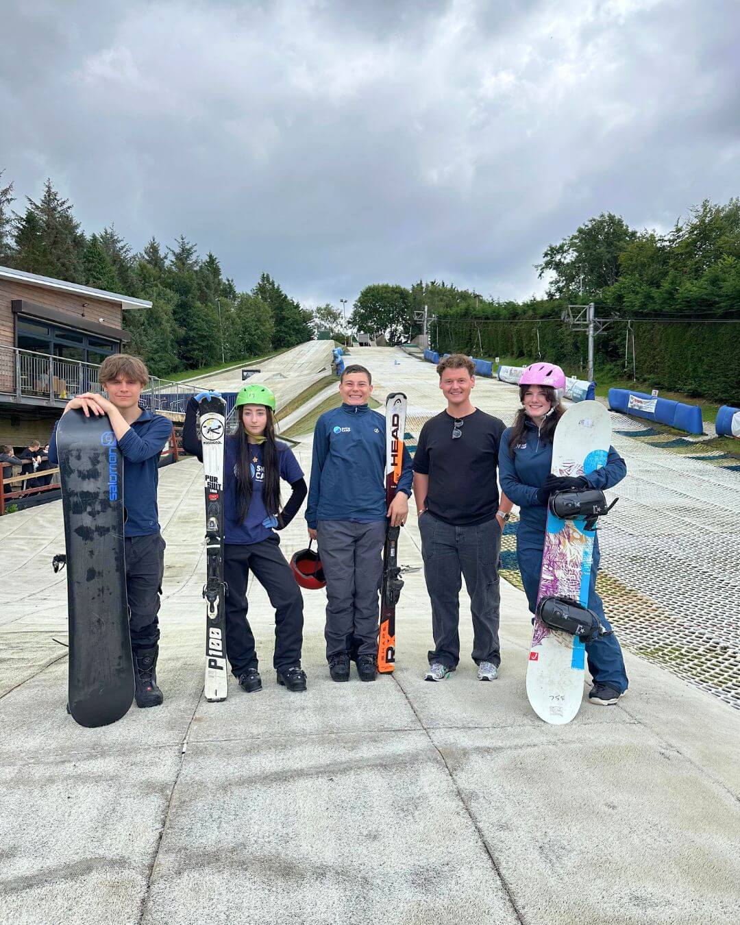 Five young people wearing ski gear stand at the bottom of a dry ski slope, holding skis and snowboards and smiling for the camera. Two are wearing helmets, indicating a training or coaching session.