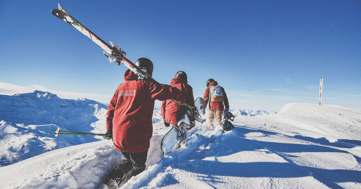 Person in a red NUCO jacket and helmet stands on a snowy ridge, watching a chairlift carry two skiers above a panoramic mountain range. The image captures the high-alpine energy of NUCO and Snow Camp’s partnership in the snowsports world.