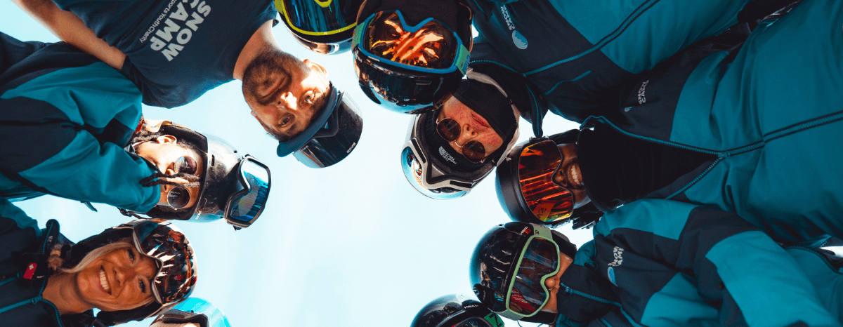 Group of Snow Camp participants and instructors form a circle and smile down at the camera, capturing team spirit and unity on the slopes.